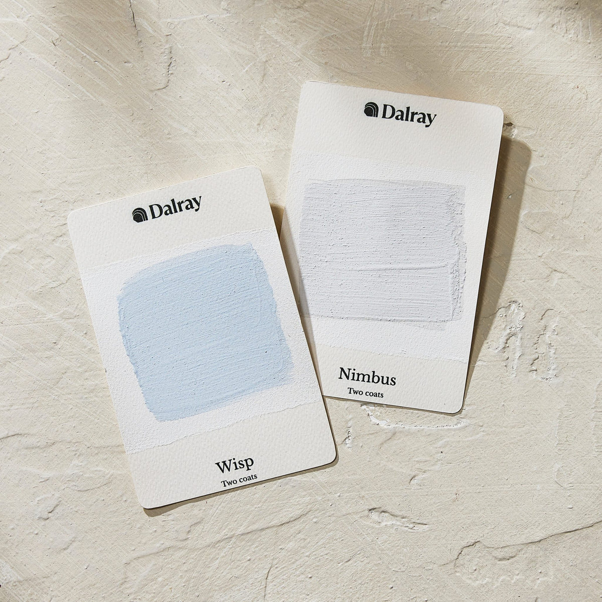 Two Dalray paint swatch cards, labeled Wisp (a pale blue swatch) and Nimbus (a very light cool gray swatch), displayed on a textured beige surface.