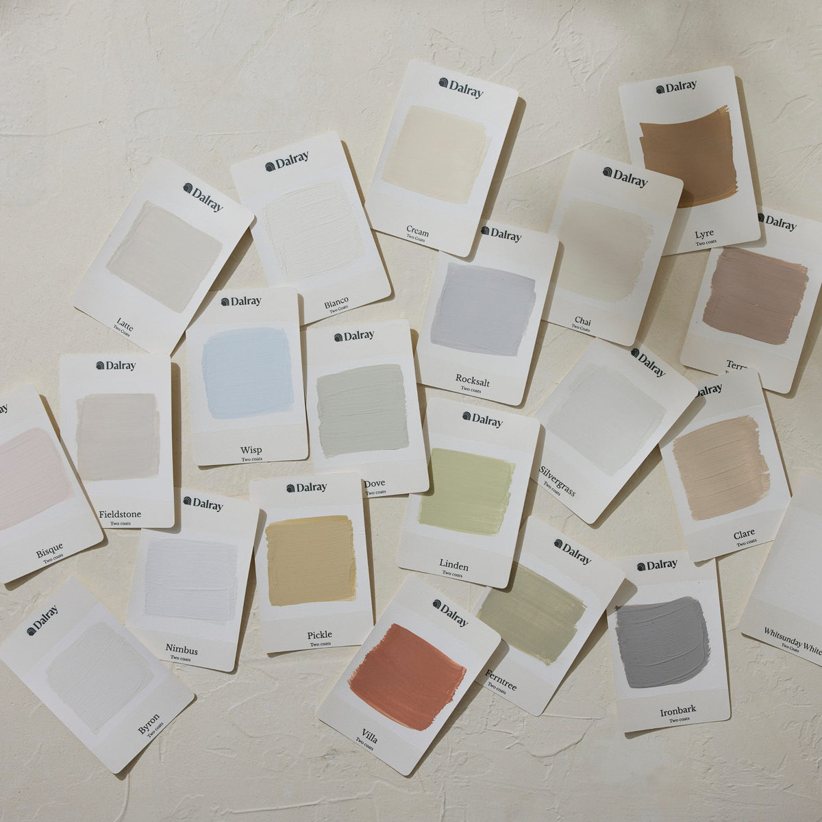 A scattered arrangement of 21 small white Dalray paint color cards on a textured off-white wall, showing multiple neutral and pastel swatches including cream, beige, gray, blue, green, pink, and brown.