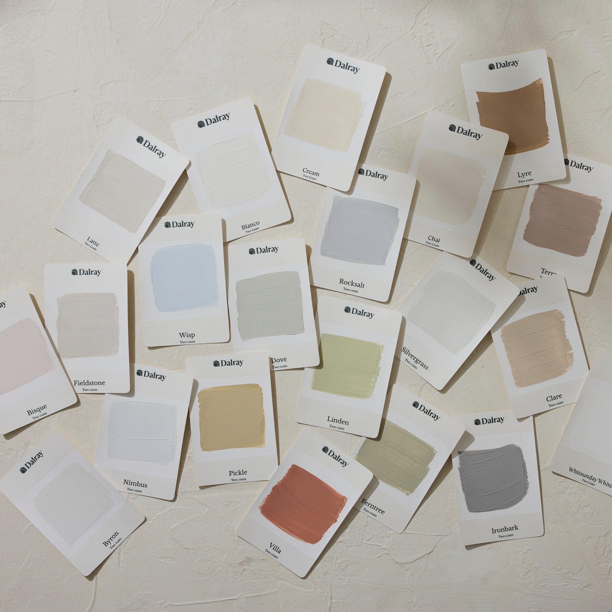 A scattered arrangement of 21 small white Dalray paint color cards on a textured off-white wall, showing multiple neutral and pastel swatches including cream, beige, gray, blue, green, pink, and brown.
