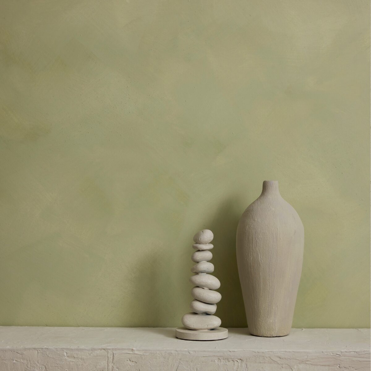 A minimalist interior scene with a textured sage green limewash wall and a white stone ledge. On the ledge sit two decorative objects: a tall matte vase on the right and a small stacked stone tower on the left.