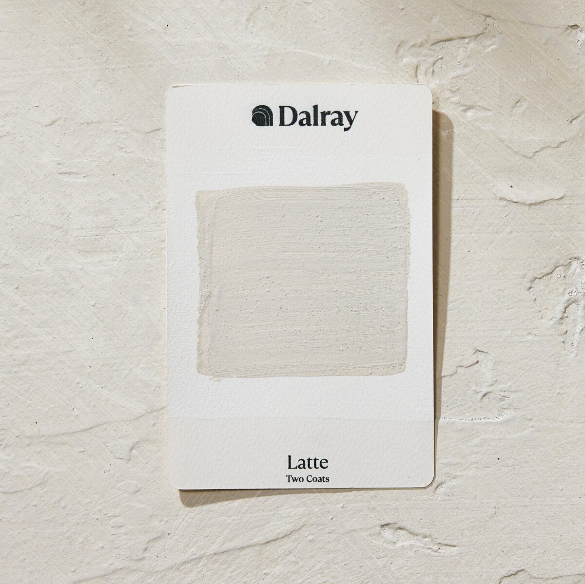 A Dalray warm paint swatch card labeled 'Latte Two Coats' showing a textured off-white neutral paint sample, placed on a textured off-white surface.