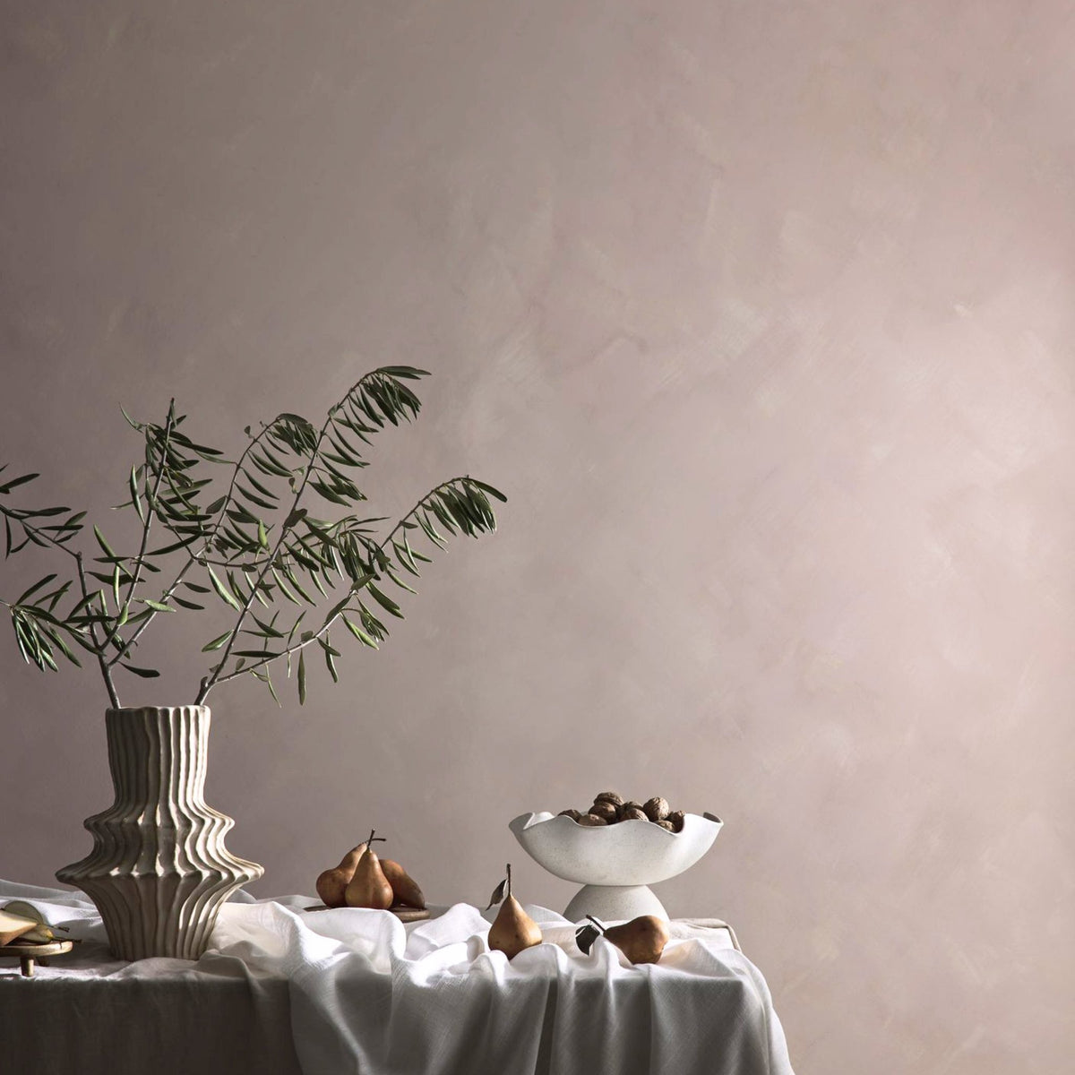 A styled interior scene with a draped table holding a ribbed ceramic vase with green stems, a white pedestal bowl with dark fruit, and a couple of pears, set against a large textured dusty pink limewash wall.