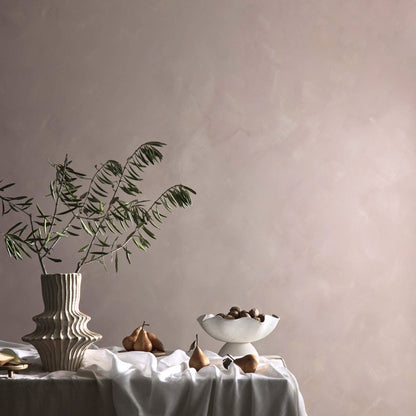A styled interior scene with a draped table holding a ribbed ceramic vase with green stems, a white pedestal bowl with dark fruit, and a couple of pears, set against a large textured dusty pink limewash wall.