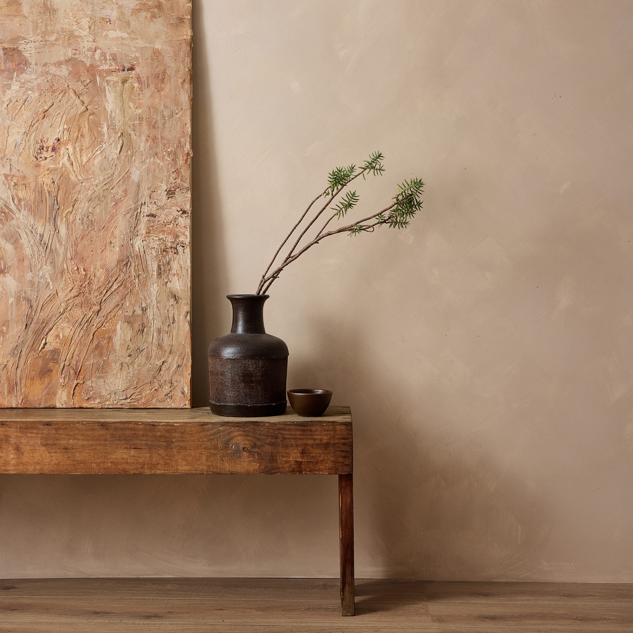 A rustic dark brown ceramic vase holding two thin branches with small green leaves, placed on a weathered wooden bench against a warm earthy beige limewash wall, with a large textured artwork partially visible on the left.