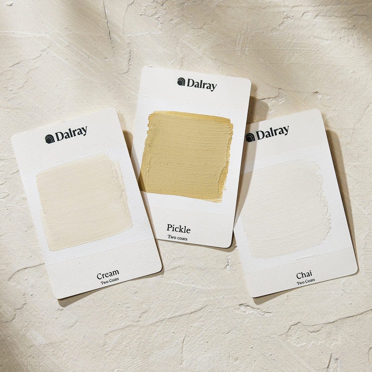 Three Dalray paint swatch cards labeled Cream, Pickle, and Chai, arranged on a textured off-white surface. The center card shows a warm beige paint swatch, while the other two show neutral beige paint swatches.