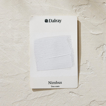 A Dalray Nimbus two-coat paint swatch card showing a textured cool blue white paint sample, placed on a light beige plaster background