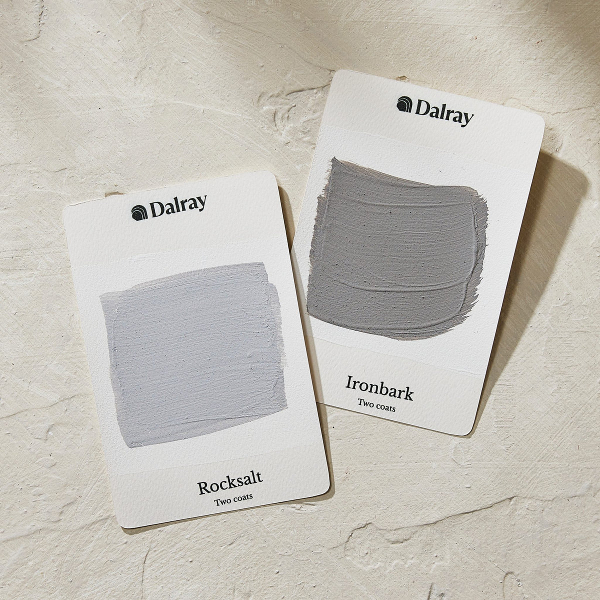 Two Dalray limewash colour cards showing textured paint swatches on a light beige stone surface. The left card is labeled 'Rocksalt' with a pale cool gray-blue swatch, and the right card is labeled 'Ironbark' with a medium warm gray swatch.