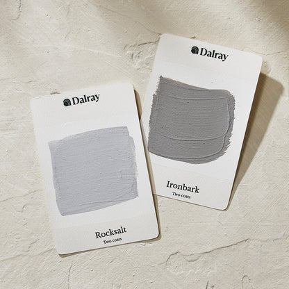 Two Dalray limewash colour cards showing textured paint swatches on a light beige stone surface. The left card is labeled 'Rocksalt' with a pale cool gray-blue swatch, and the right card is labeled 'Ironbark' with a medium warm gray swatch.