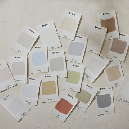 A scattered arrangement of 21 small white Dalray paint color cards on a textured off-white wall, showing multiple neutral and pastel swatches including cream, beige, gray, blue, green, pink, and brown.
