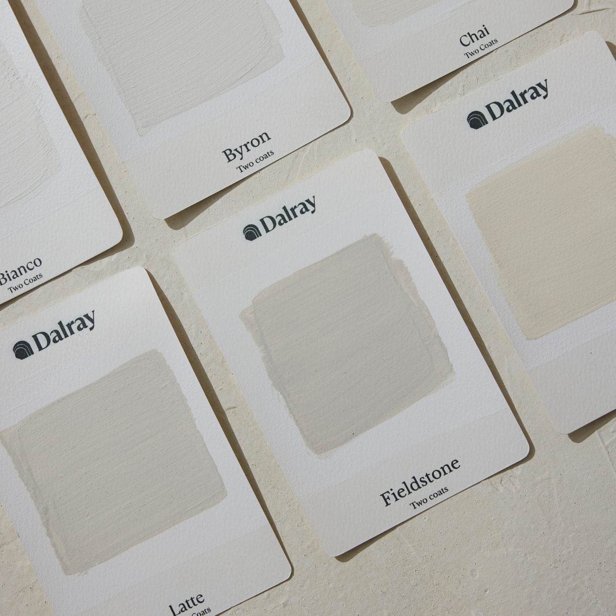 A close-up of several Dalray limewash paint swatch cards arranged on a light textured surface. The visible cards show painted color samples and printed names including Byron, Chai, Fieldstone, and Latte.