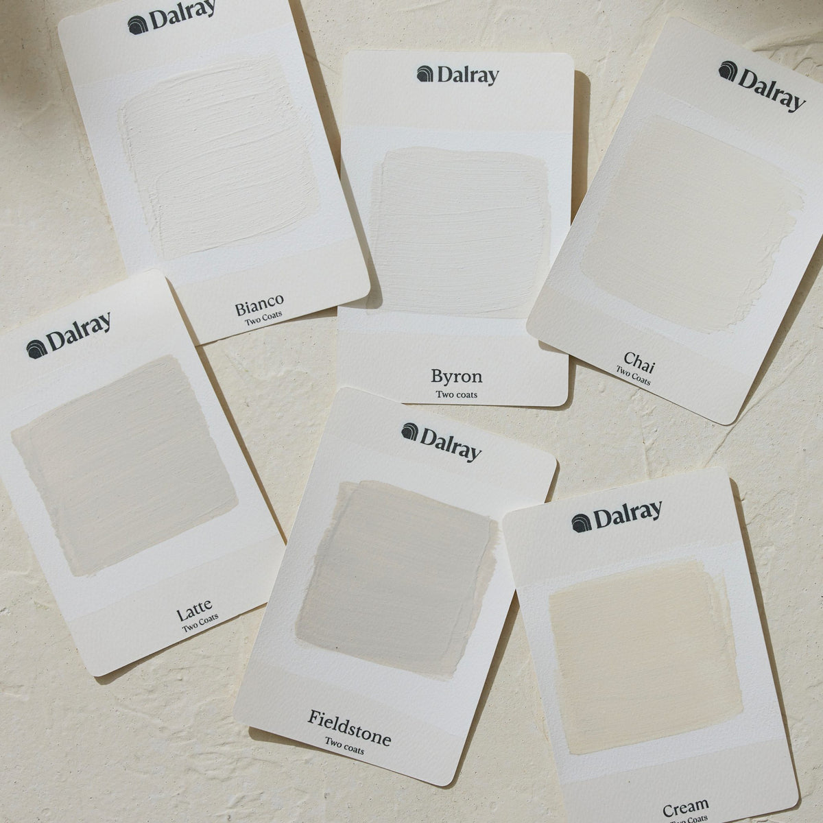 Six Dalray paint A6 swatch cards arranged on a textured off-white surface, showing different limewash paint warm off-white and cream shades labeled Bianco, Byron, Chai, Latte, Fieldstone, and Cream.