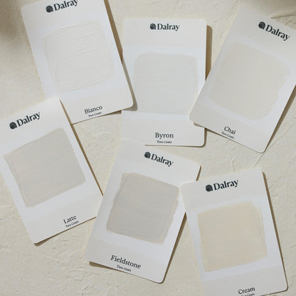 Six Dalray paint A6 swatch cards arranged on a textured off-white surface, showing different limewash paint warm off-white and cream shades labeled Bianco, Byron, Chai, Latte, Fieldstone, and Cream.