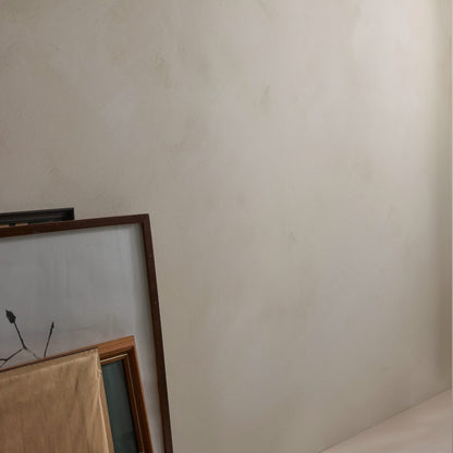 A close-up of a textured, warm off-white limewash wall in Dalray limewash colour Chai with the corner of a wooden-framed artwork and a beige fabric surface visible in the lower-left corner.
