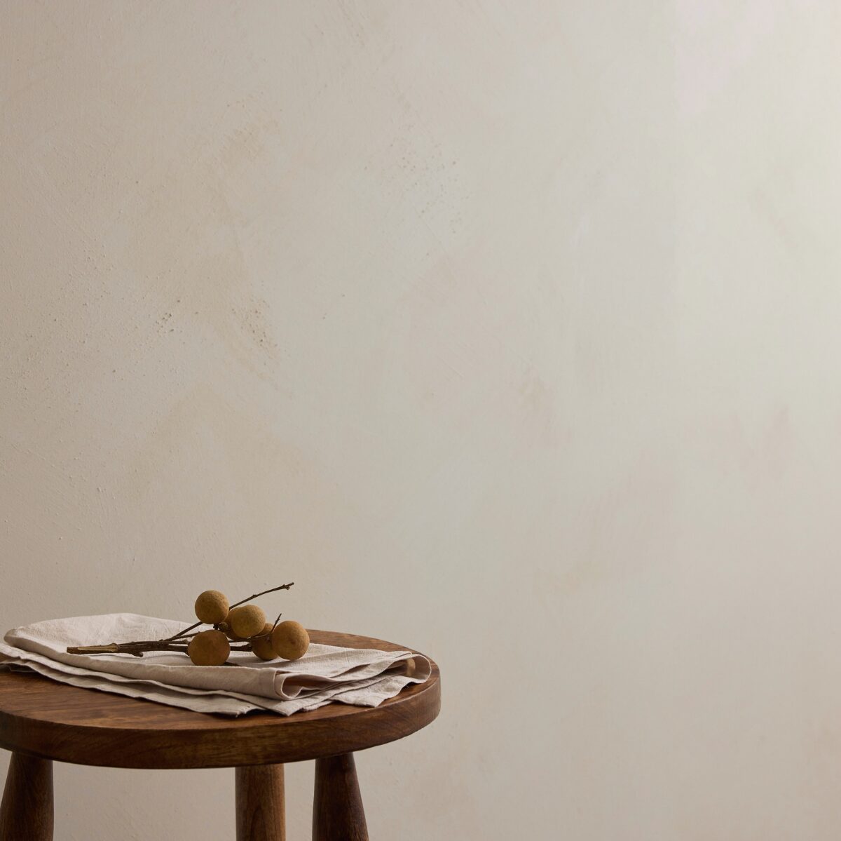 A round wooden stool with a folded beige linen cloth and a small bunch of brown fruit on top, set against a plain textured off-white Chai Limewash wall