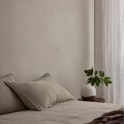 A styled bedroom corner with a bed dressed in neutral beige bedding, matching pillows, a small wooden side table, a speckled white ceramic vase with green leaves, and sheer white curtains by a softly lit window. Dalray limewash paint on back wall in colour Latte - textured wall