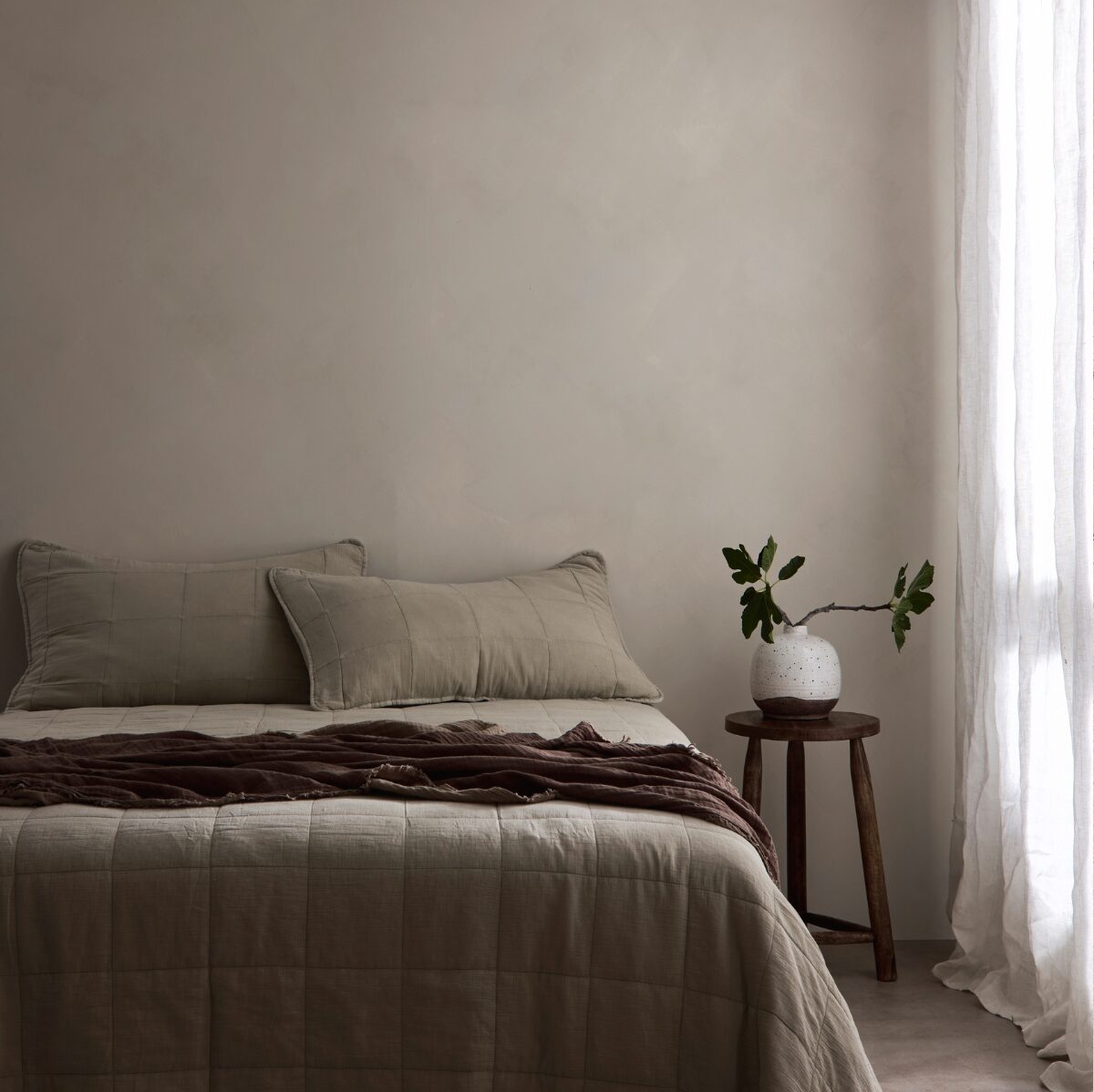 A neatly made bed in a minimalist bedroom setting, featuring beige quilted bedding, two matching pillows, a dark brown throw blanket, a small wooden side table with a white vase of green stems, and sheer white curtains by a window. Dalray australia limewash paint in colour Latte is on the wall in the background.