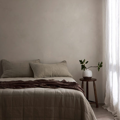 A neatly made bed in a minimalist bedroom setting, featuring beige quilted bedding, two matching pillows, a dark brown throw blanket, a small wooden side table with a white vase of green stems, and sheer white curtains by a window. Dalray australia limewash paint in colour Latte is on the wall in the background.