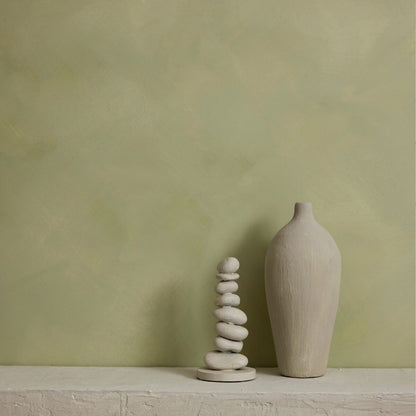 A minimalist interior scene with a textured sage green limewash wall and a white stone ledge. On the ledge sit two decorative objects: a tall matte vase on the right and a small stacked stone tower on the left.