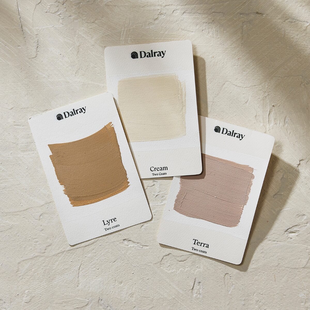 Three Dalray limewash paint swatch cards showing cream, Lyre, and Terra shades, arranged on a textured beige surface.