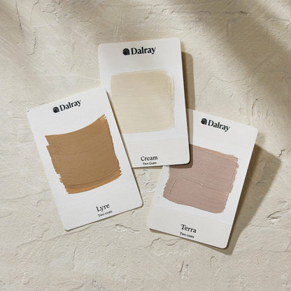 Three Dalray limewash paint swatch cards showing cream, Lyre, and Terra shades, arranged on a textured beige surface.