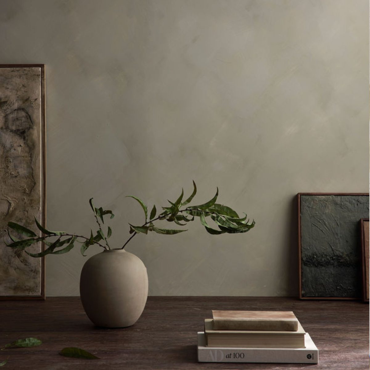 A minimalist interior scene with a matte beige ceramic vase holding green leafy stems on a dark wooden table, with stacked books and framed wall art in the background. Dark forest moody green limewash in the background creating a textured wall.
