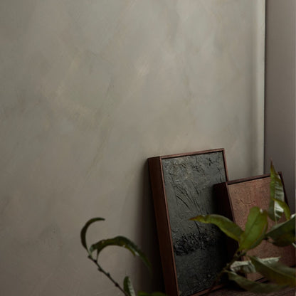 Two framed textured art prints leaning against a neutral green limewash wall, with green plant leaves in the foreground.